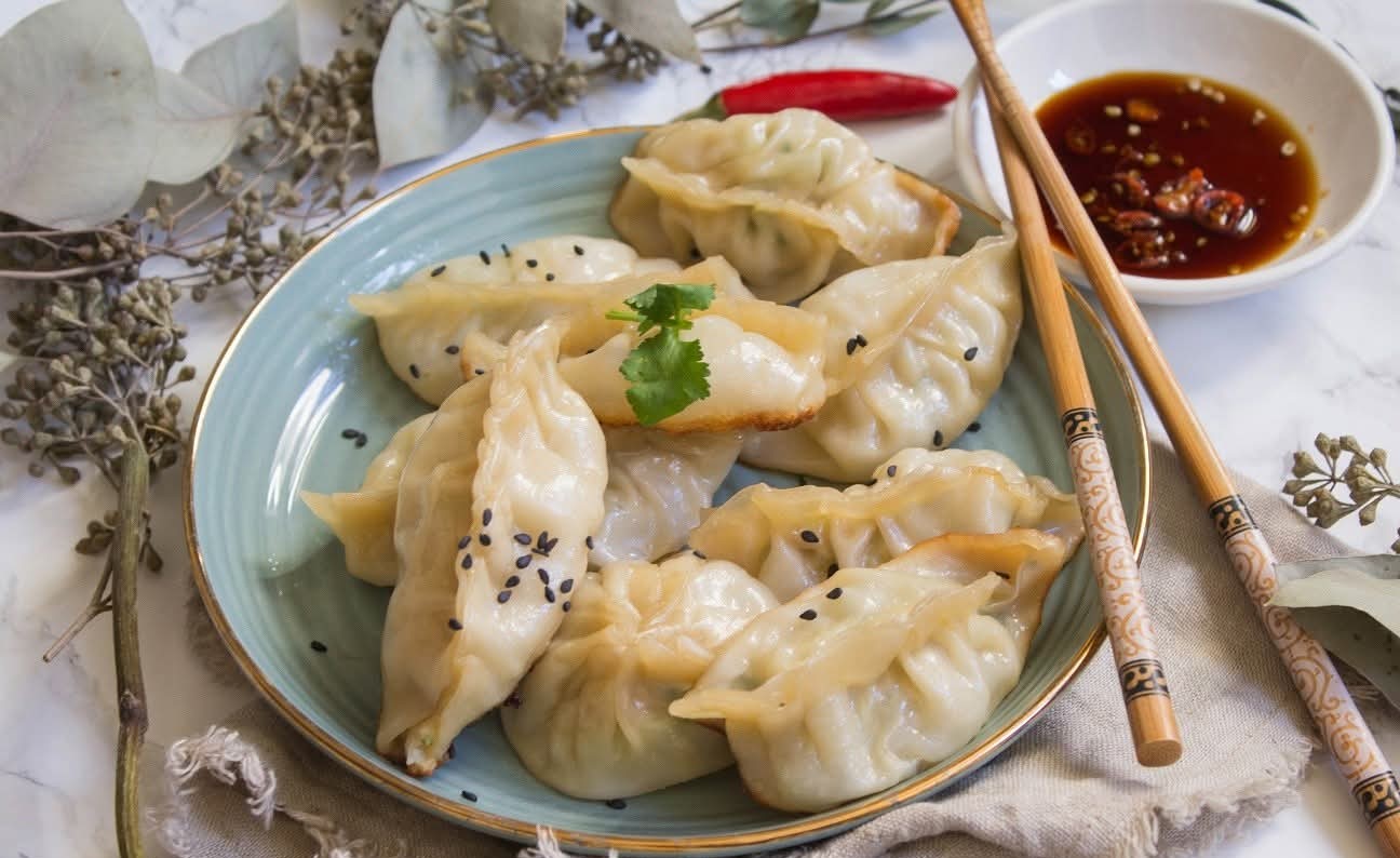 Steamed dumplings with pleated edges, garnished with cilantro and sesame seeds, served with chilli dipping sauce.