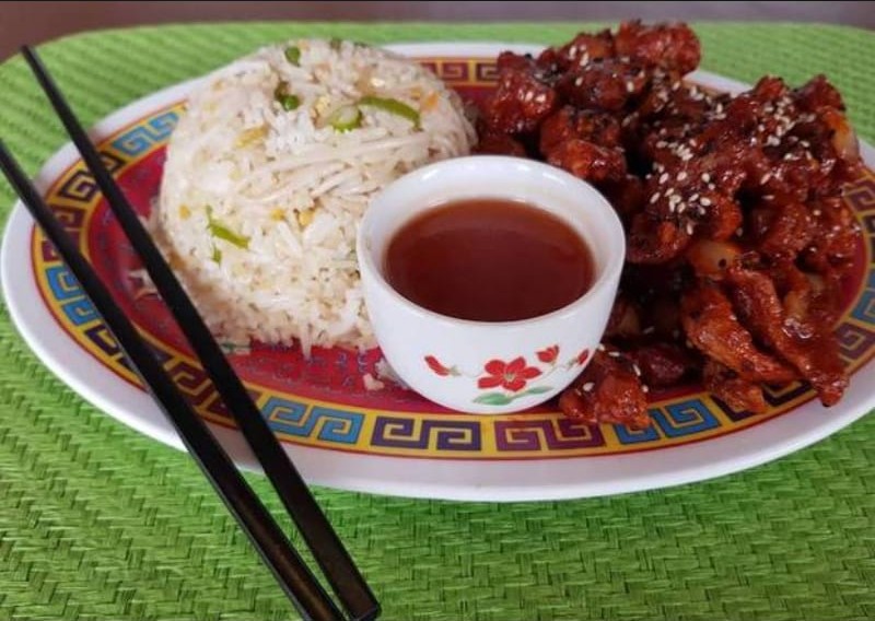Crispy glazed chicken topped with white sesame seeds, served with fried rice and a dipping sauce on a patterned plate.